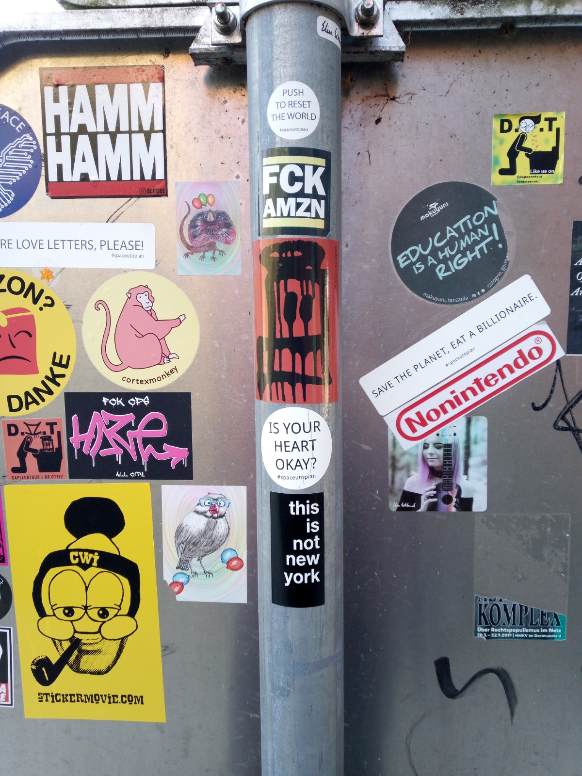 This image shows a round, white, minimalist sticker from the street artist @Space_Utopian with the text "Is your heart okay?" on a grey pole somewhere in the streets of Ljubljana.